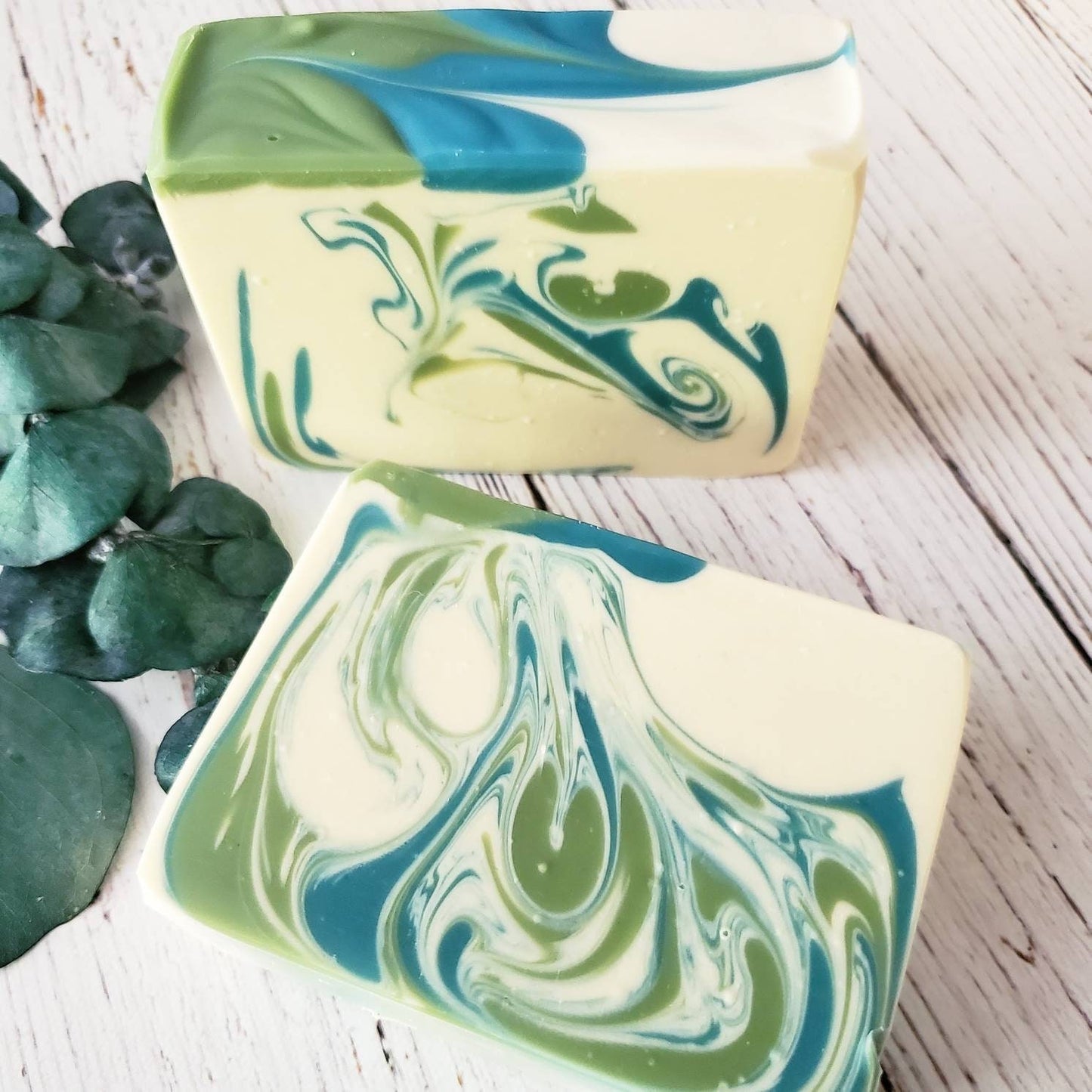 Two bars of soap with green and blue swirls placed on a white wooden surface next to eucalyptus leaves.